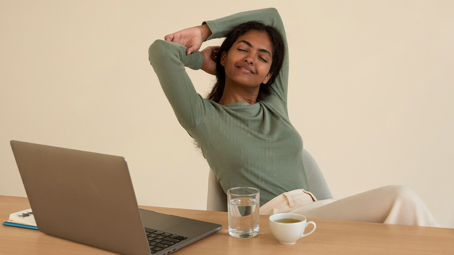 A South Asian woman leaning back at her desk doing a relaxed stretch during an afternoon screen break, with a glass of water and a cup of herbal tea.