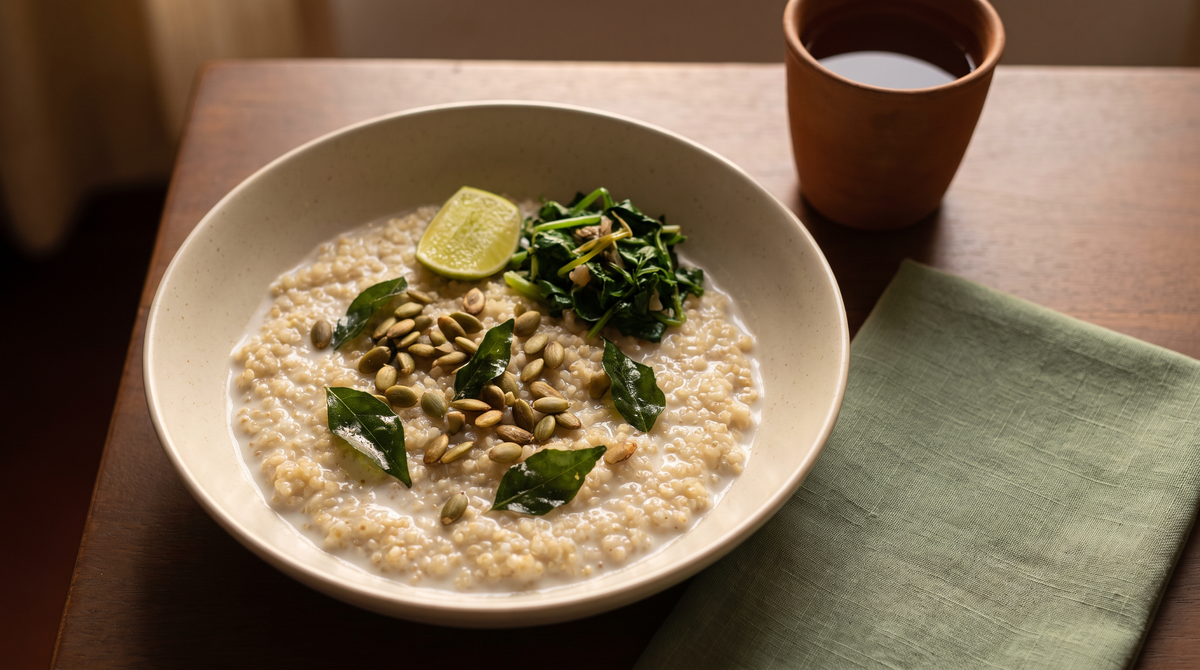 A ceramic bowl of creamy coconut quinoa topped with toasted pumpkin seeds, curry leaves, wilted greens and a lime wedge on a dark wooden table in evening light.