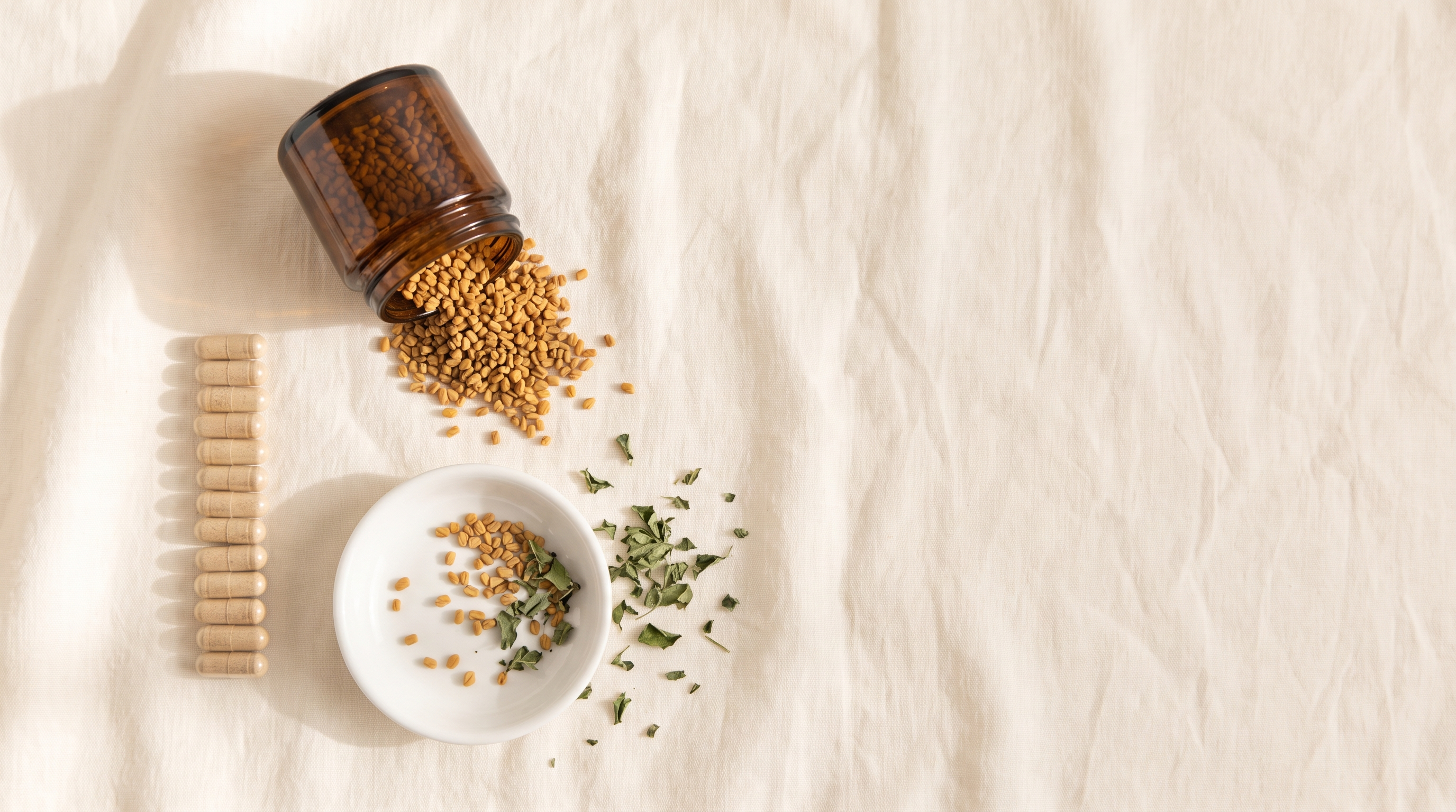Fenugreek seeds spilling from a plain amber jar beside a row of beige capsules on cream linen