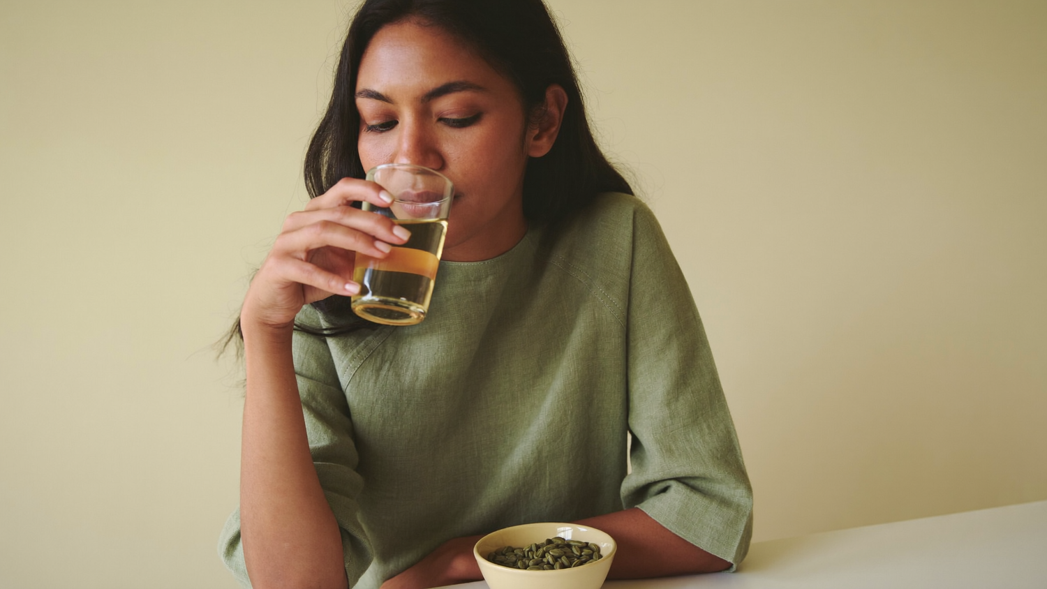 A South Asian woman in a sage green top sips a pale golden infusion beside a small bowl of raw unroasted green coffee beans at a sunlit table.