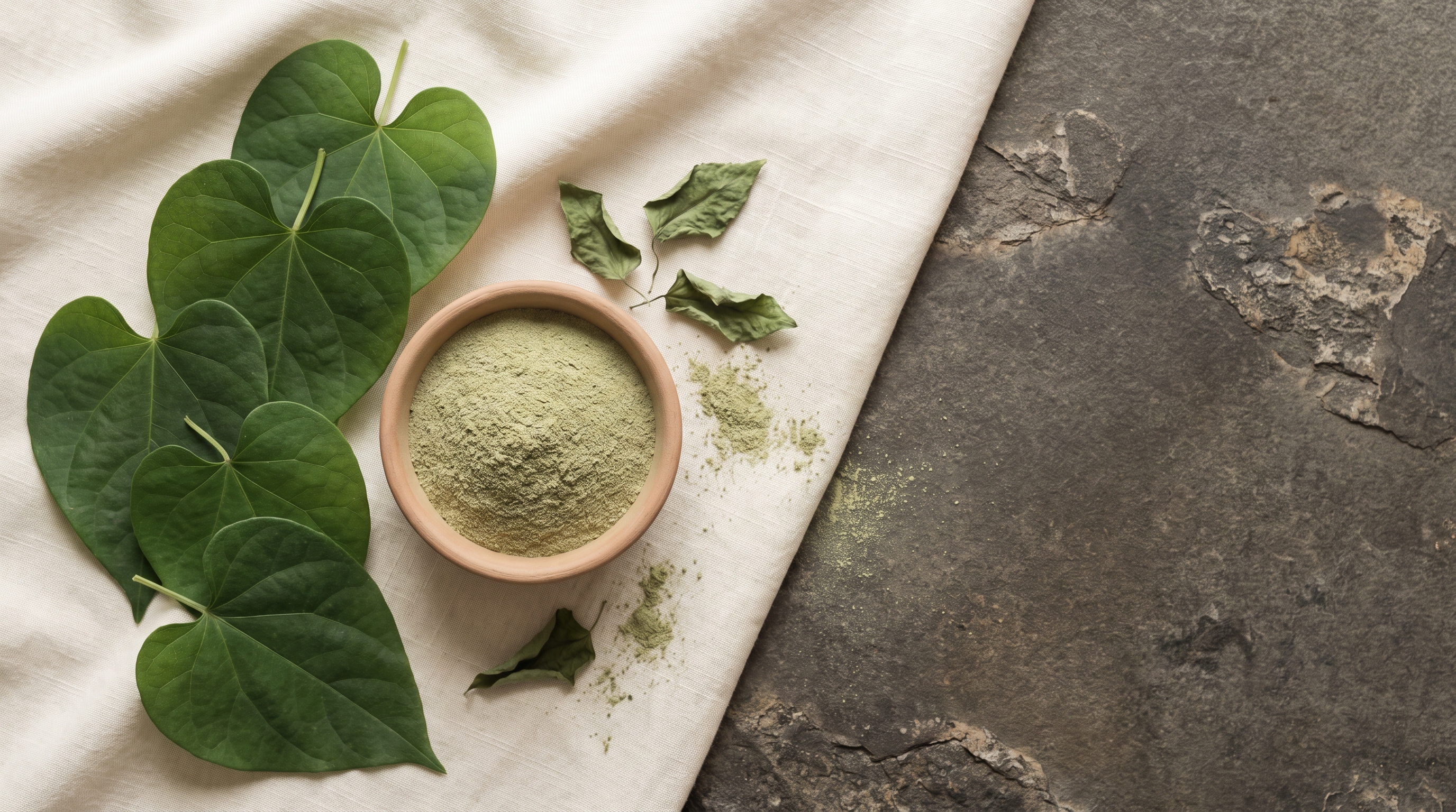 Fresh green vine leaves and a plain clay bowl of milled green leaf powder on cream linen over a dark stone table