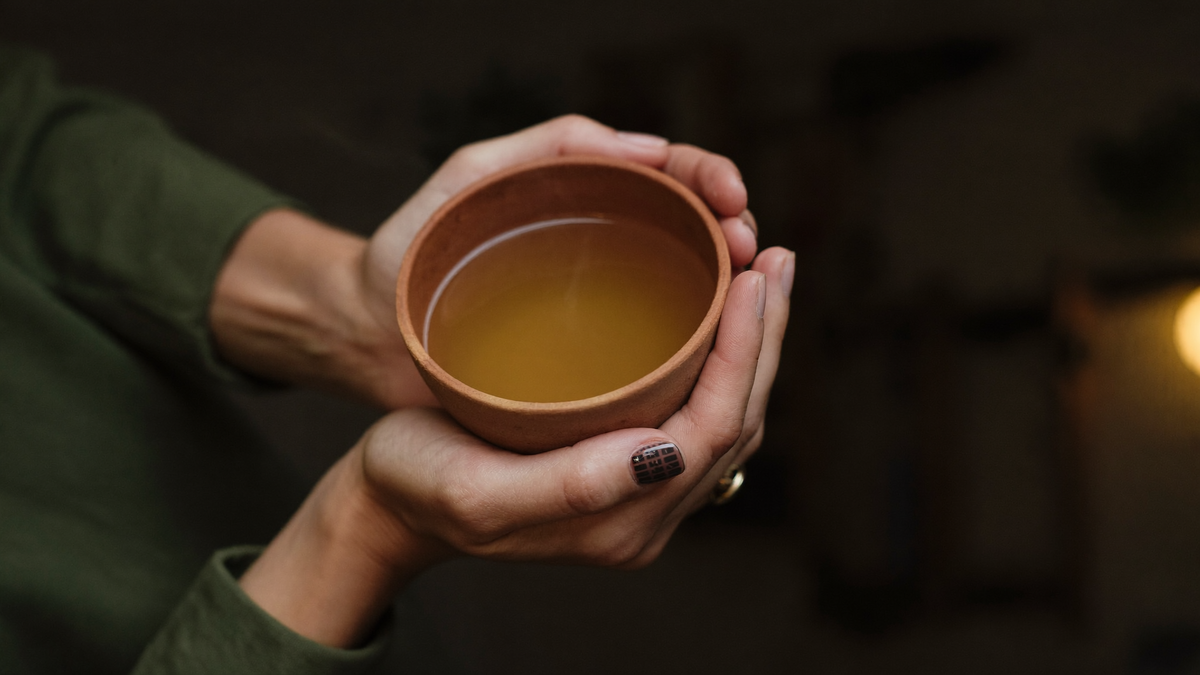 Two hands cupping a small warm clay cup of golden herbal tonic in soft evening light