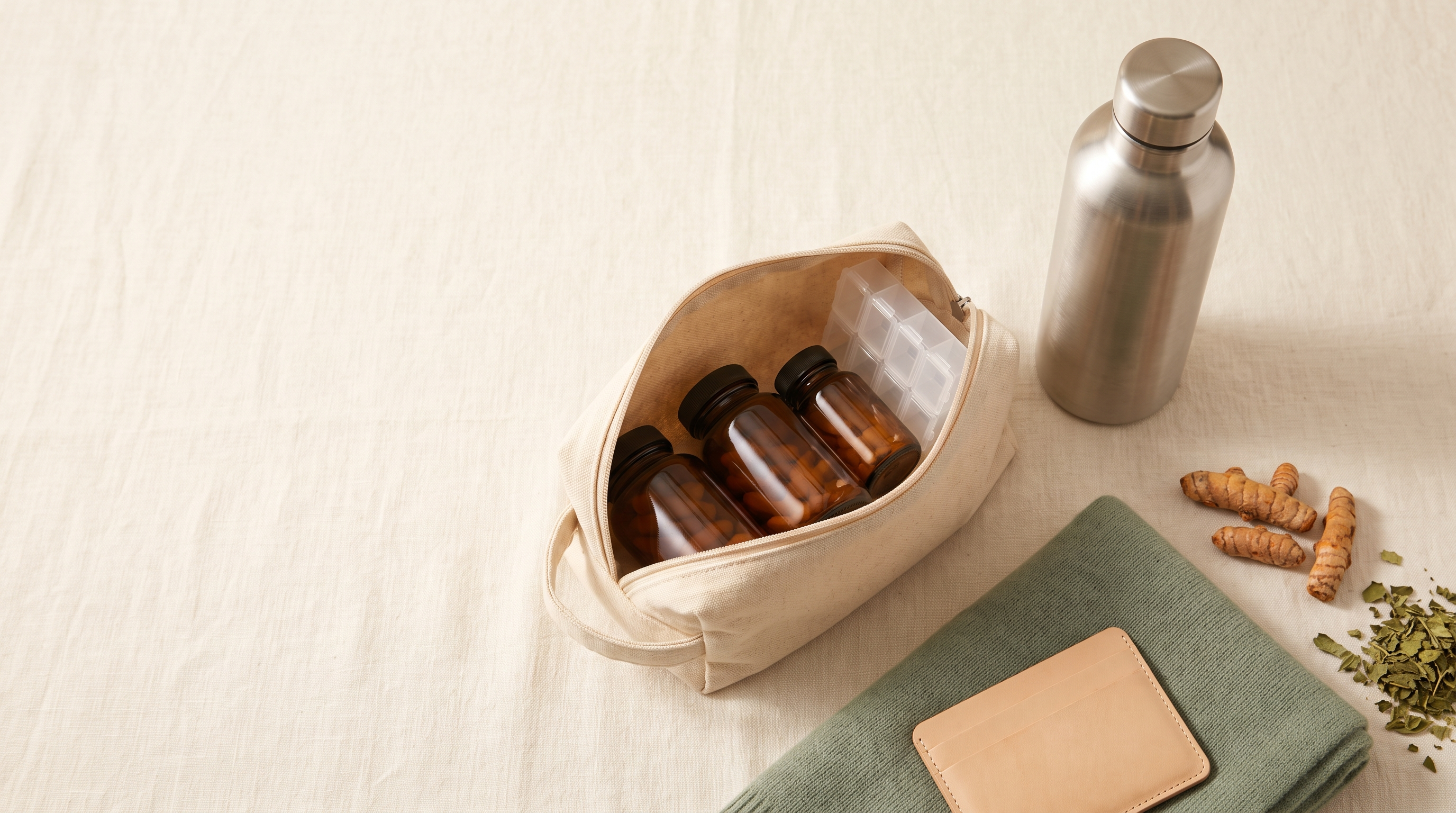 A canvas travel pouch holding unlabelled amber supplement bottles and a pill organiser, beside a steel water bottle, turmeric root and dried leaves on pale linen