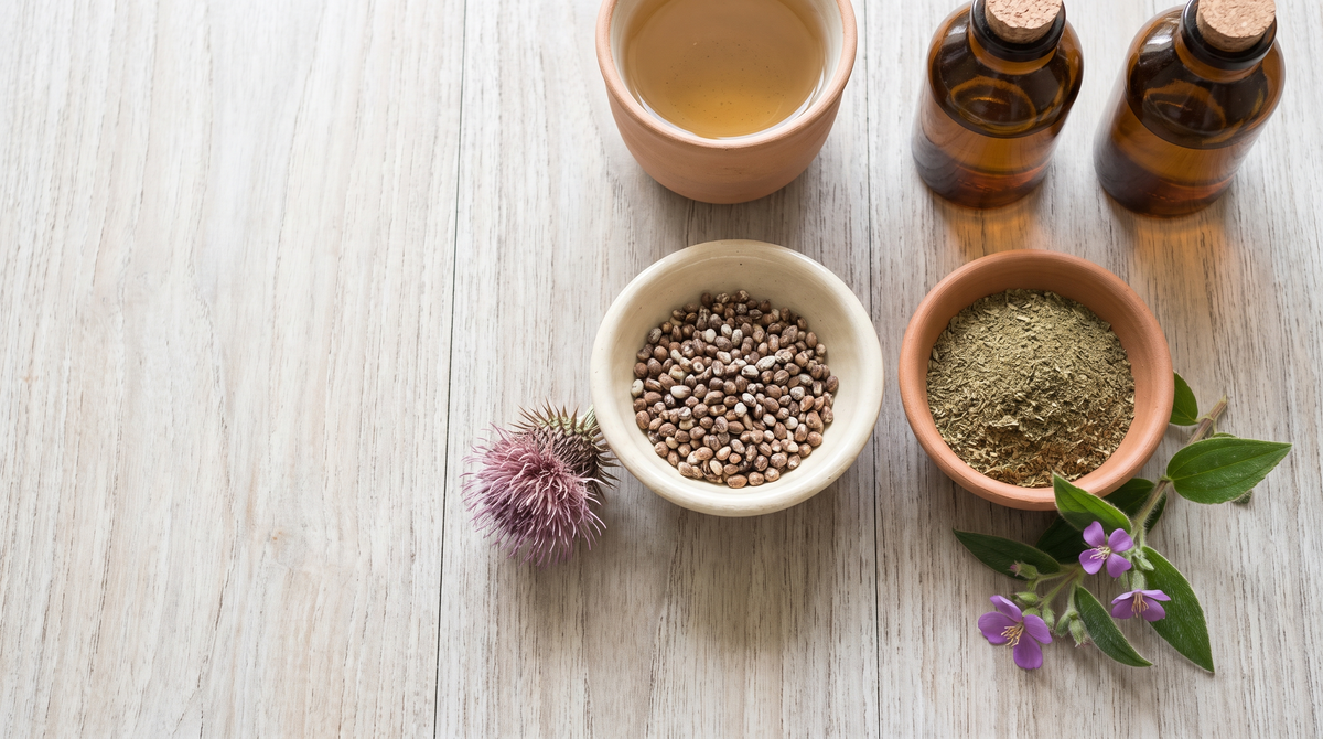 Overhead still life of a bowl of milk thistle seeds with a purple thistle flower beside a bowl of green Heenbovitiya leaf powder and a flowering sprig, with a clay cup of herbal tea and two plain amber bottles