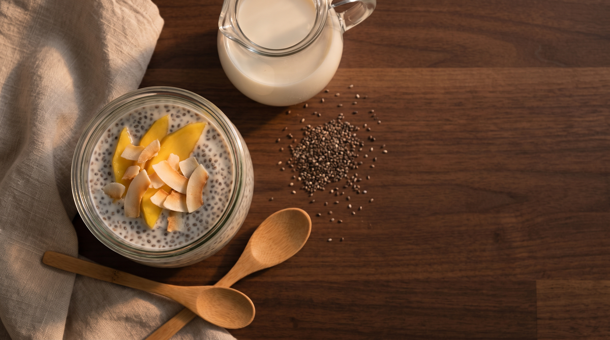 A glass jar of overnight-soaked chia seeds in coconut milk topped with mango and coconut flakes, beside dry chia seeds and wooden spoons on a dark wood counter