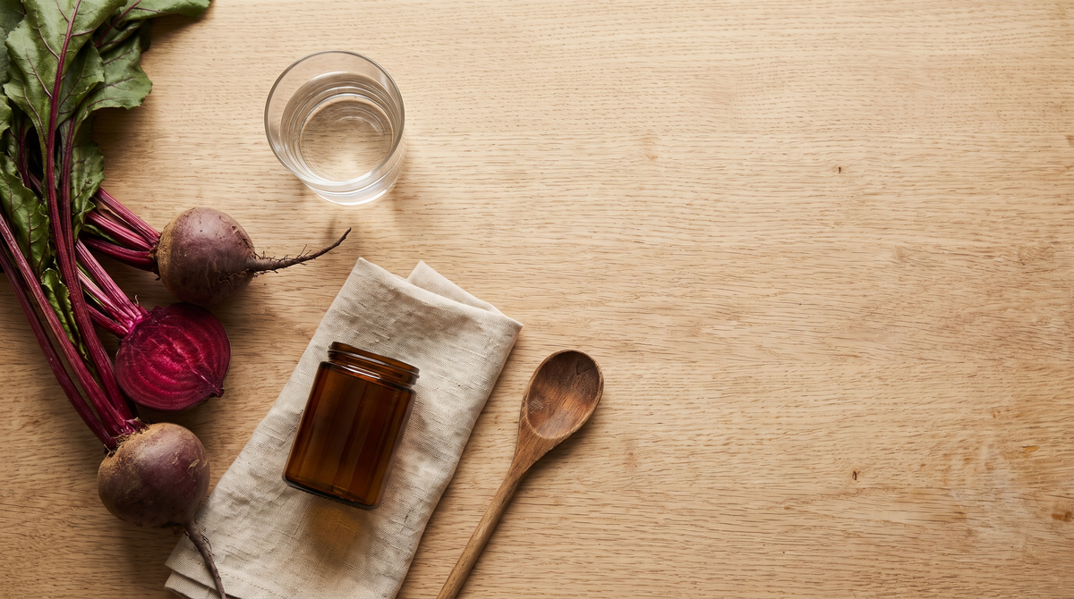 Whole beetroots, a halved beetroot, a glass of water and an unlabeled amber jar on a light oak table