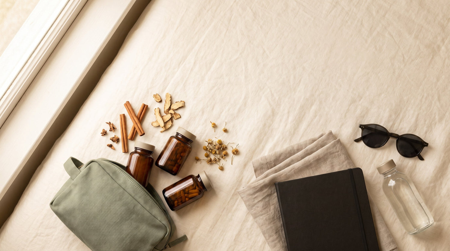 A flat-lay of a travellers natural wellness kit: a sage green pouch, three unlabeled amber capsule bottles, dried cinnamon and herbs, a notebook, sunglasses, and a water bottle on a cream linen surface.