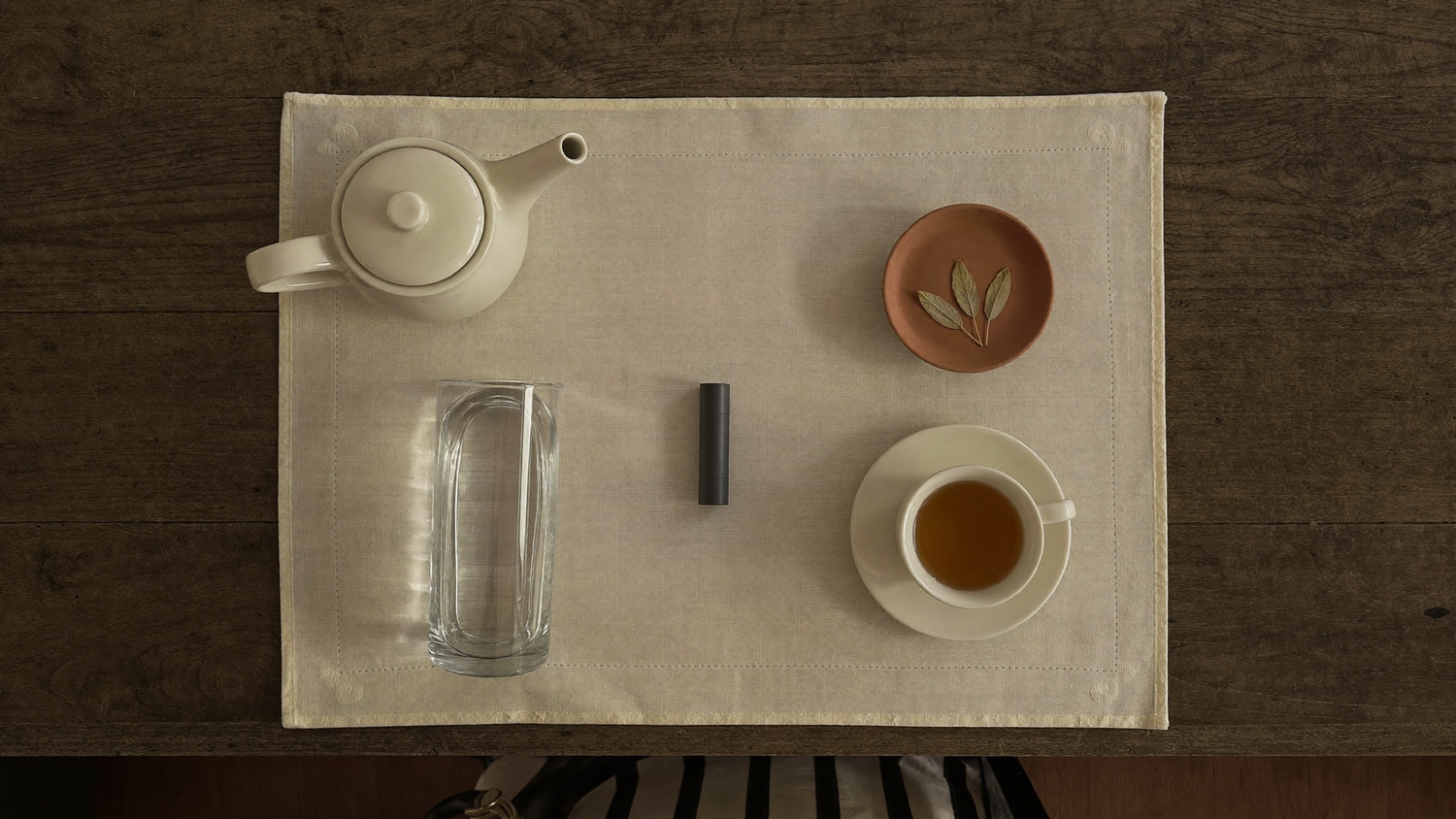 Overhead flat-lay of an evening cleanse ritual setup with cream teapot, glass of water, capsule, and amber herbal tea on plain linen.