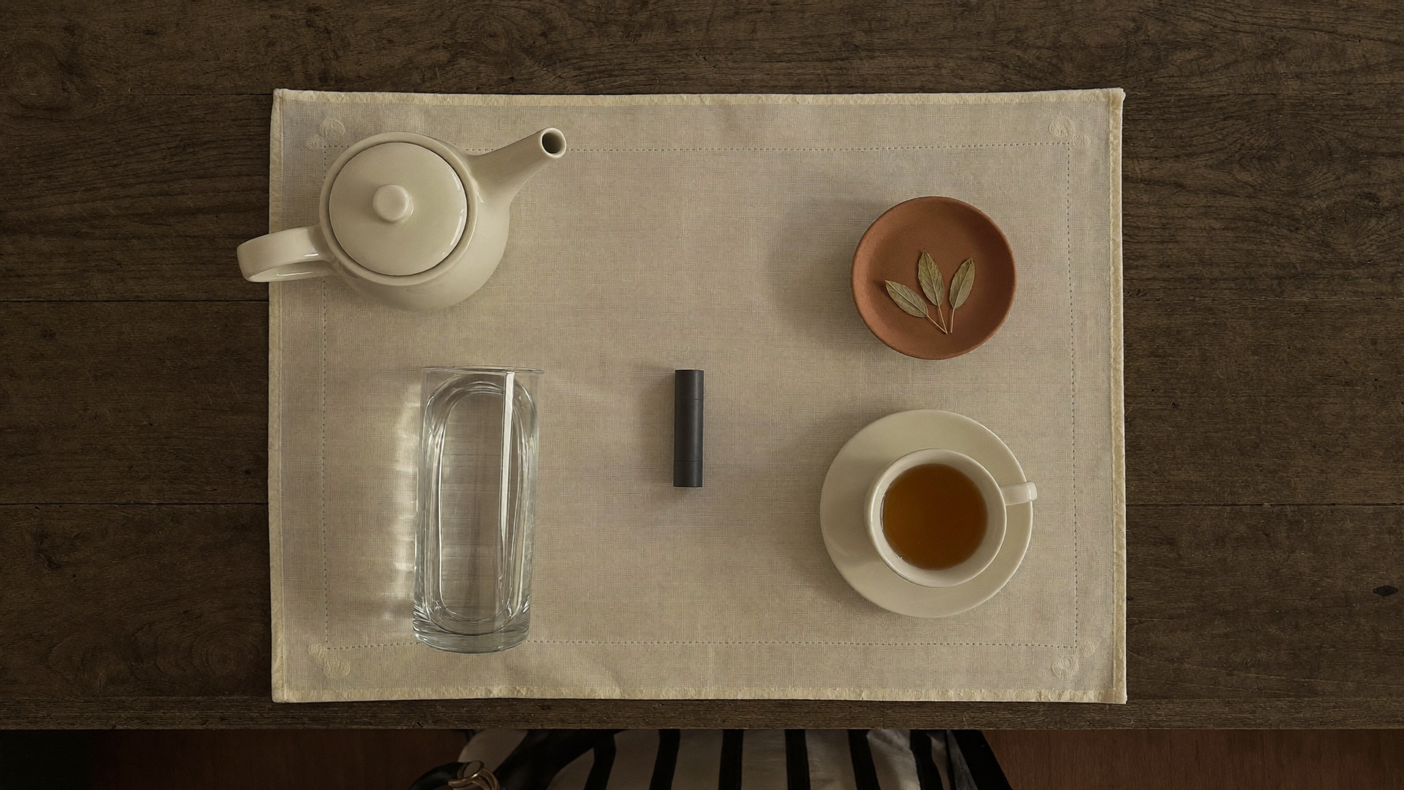 Overhead flat-lay of an evening cleanse ritual setup with cream teapot, glass of water, capsule, and amber herbal tea on plain linen.