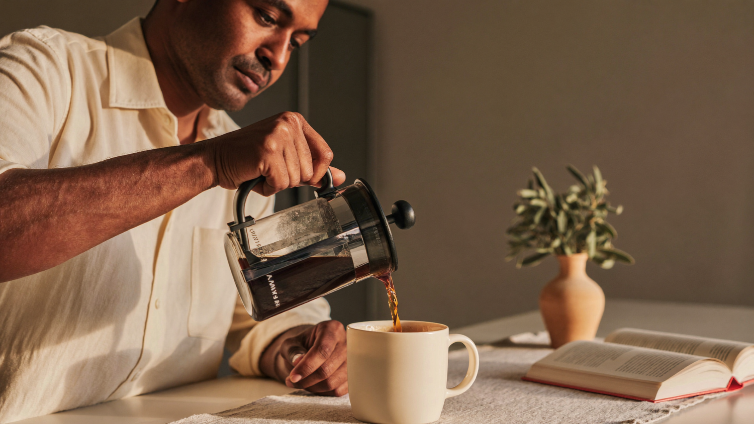 A South Asian man pouring coffee from a French press into a cream ceramic mug at a sunlit kitchen counter, modern wellness lifestyle aesthetic.