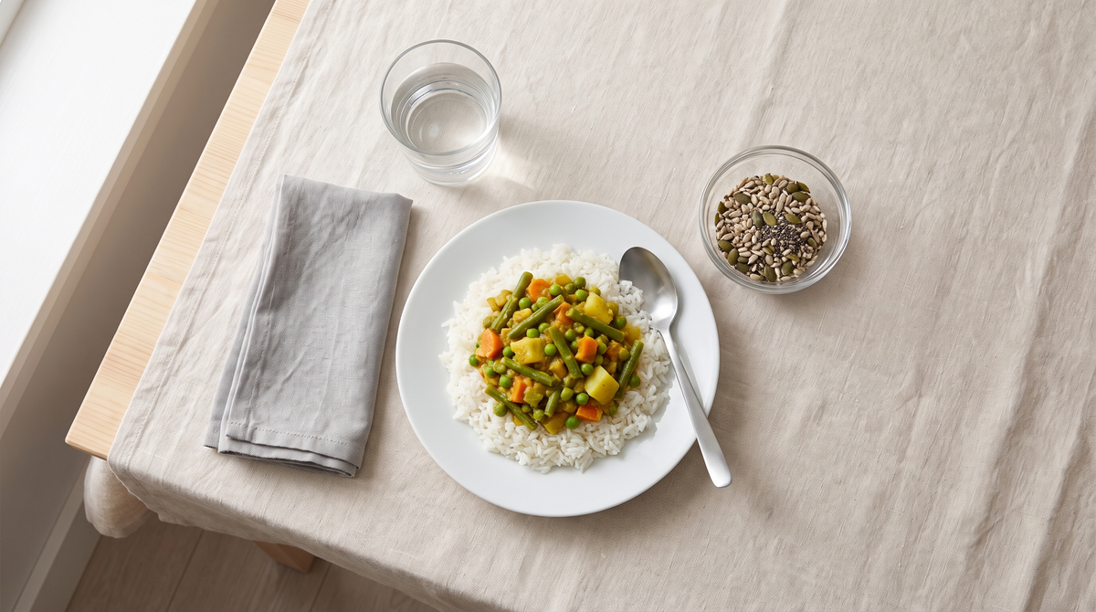 Overhead view of a rice and vegetable curry lunch on a linen tablecloth, with a small glass bowl of mixed seeds and a glass of water