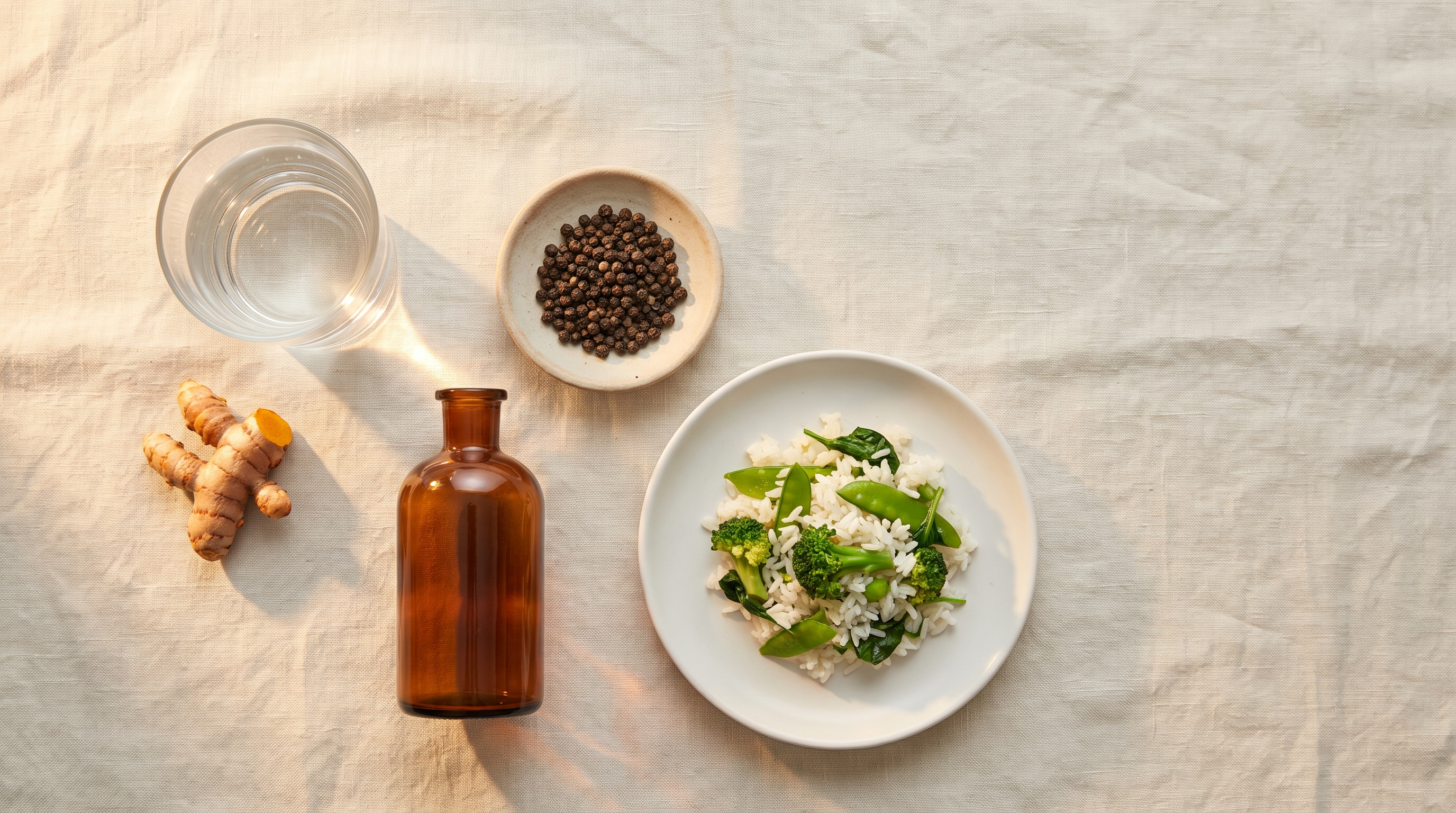 Overhead flat lay of a glass of water, fresh turmeric root, black peppercorns and a small plate of rice with green vegetables on pale linen