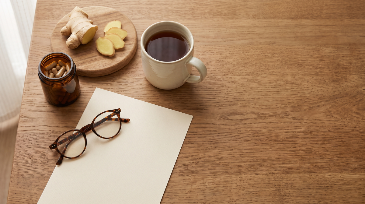 Overhead view of an oak table with a jar of capsules, fresh ginger, a cup of tea and reading glasses on a blank sheet of paper