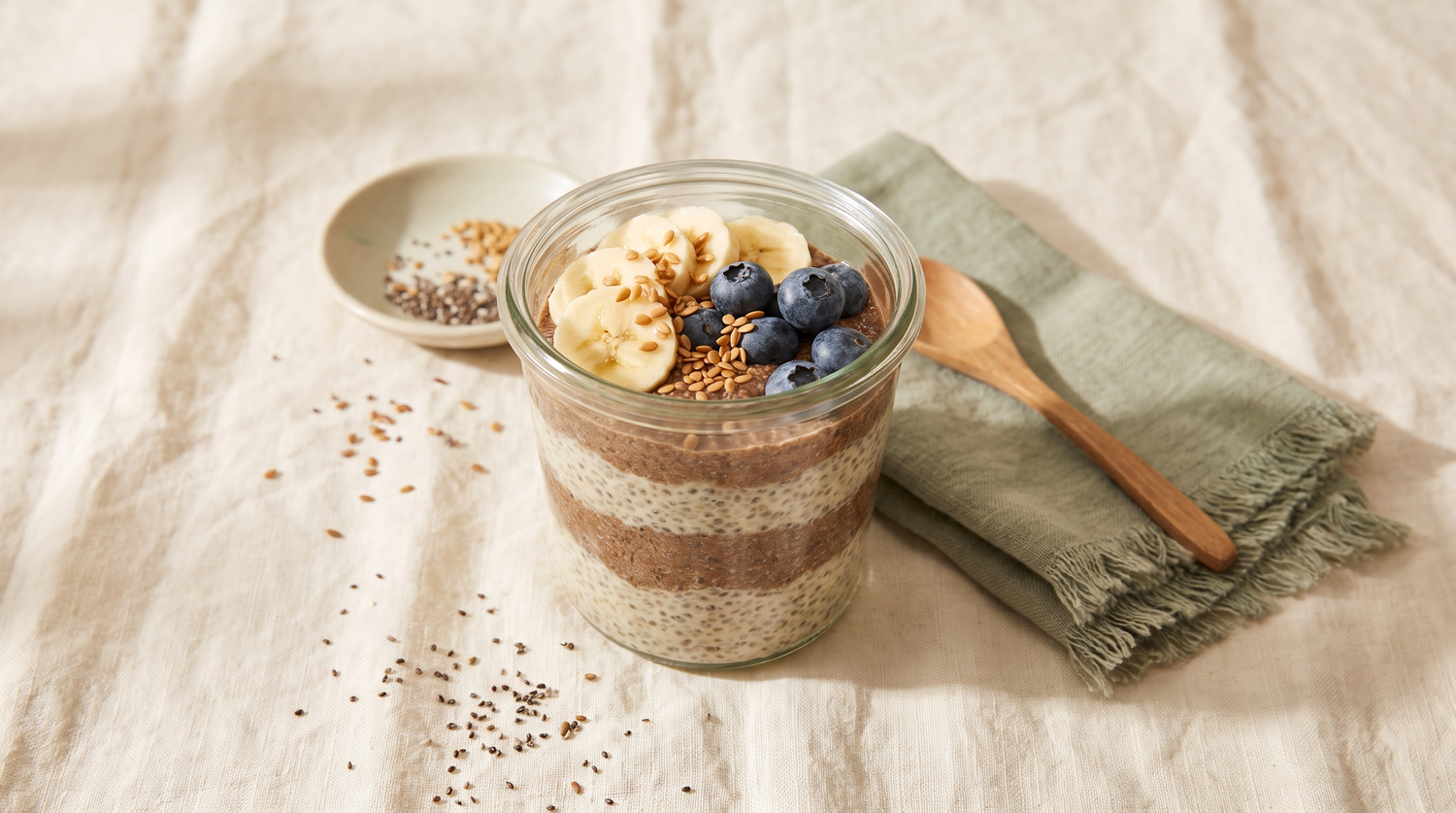 A layered flax and chia overnight bowl in a glass jar topped with banana, blueberries and flax seeds on cream linen