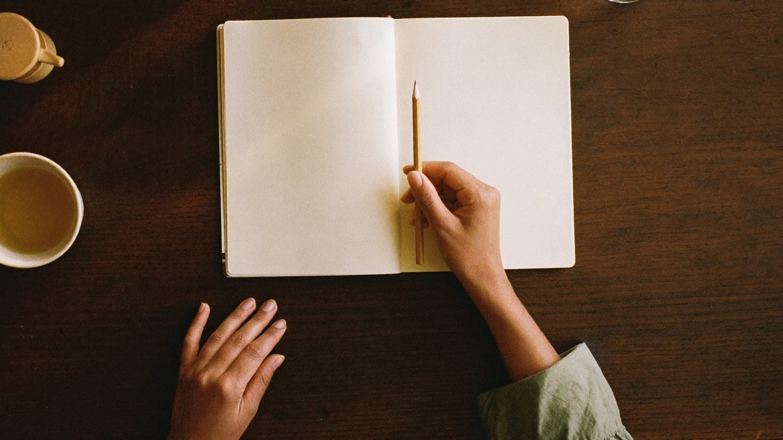 Overhead view of a blank open notebook, a pencil, a cup of herbal tea and a womans hands resting on a dark wooden table in warm evening light