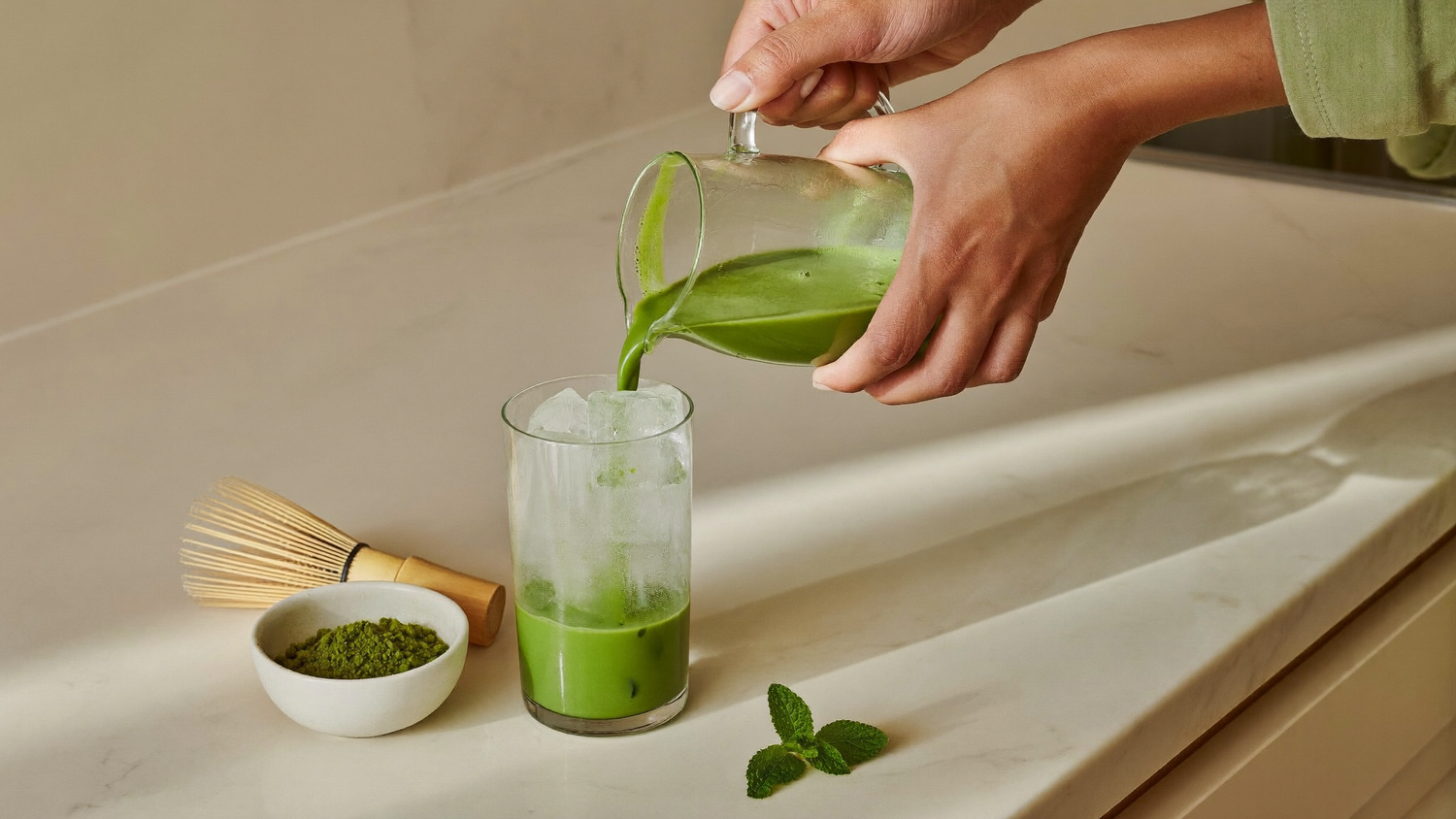 Two hands pouring bright green iced ceremonial matcha from a glass jug over a tall glass of ice, with a bamboo whisk, a bowl of matcha powder and a mint sprig on a cream marble counter.
