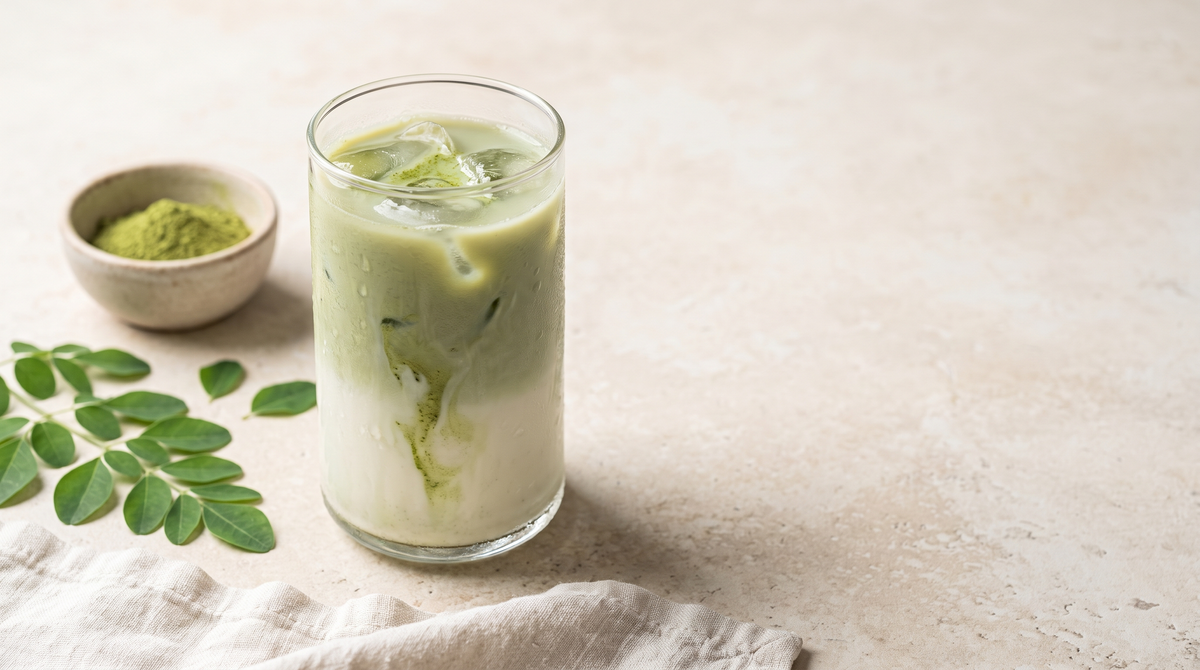 A tall glass of iced green latte with ice, a small bowl of green moringa leaf powder and fresh moringa leaves on a pale stone counter