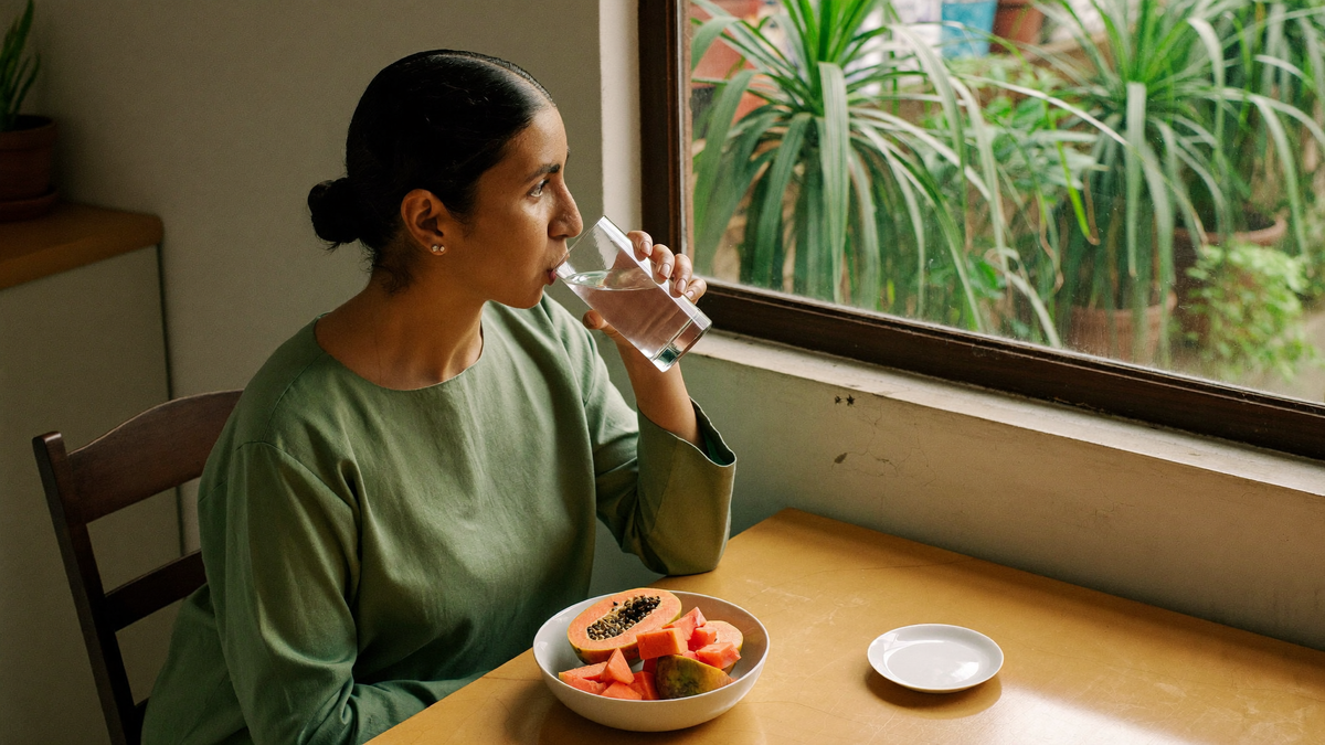 A woman in a sage green top drinks a glass of water at a wooden breakfast table beside a sunlit window, part of a calm morning focus ritual.