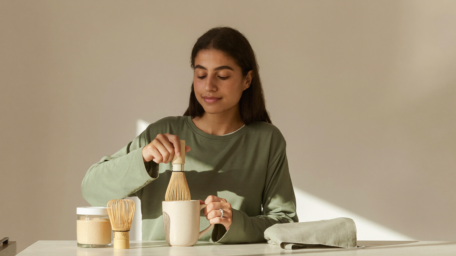 A South Asian woman whisking a warm frothy mushroom latte in a plain ceramic mug at a sunlit kitchen counter in the early morning.