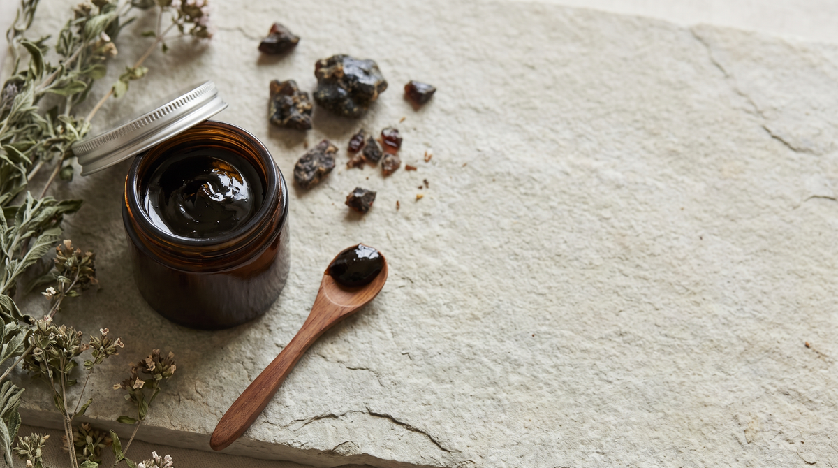 Open amber glass jar of dark mineral resin with a wooden spoon and resin fragments on a pale stone surface