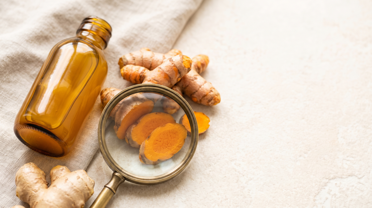 A magnifying glass held over sliced fresh turmeric beside an unlabelled amber glass wellness shot and fresh ginger root
