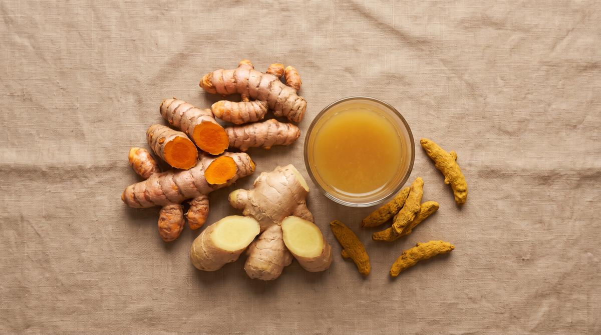 Overhead flat lay of fresh turmeric and ginger rhizomes cut open beside a small glass of golden liquid and dried turmeric fingers on linen