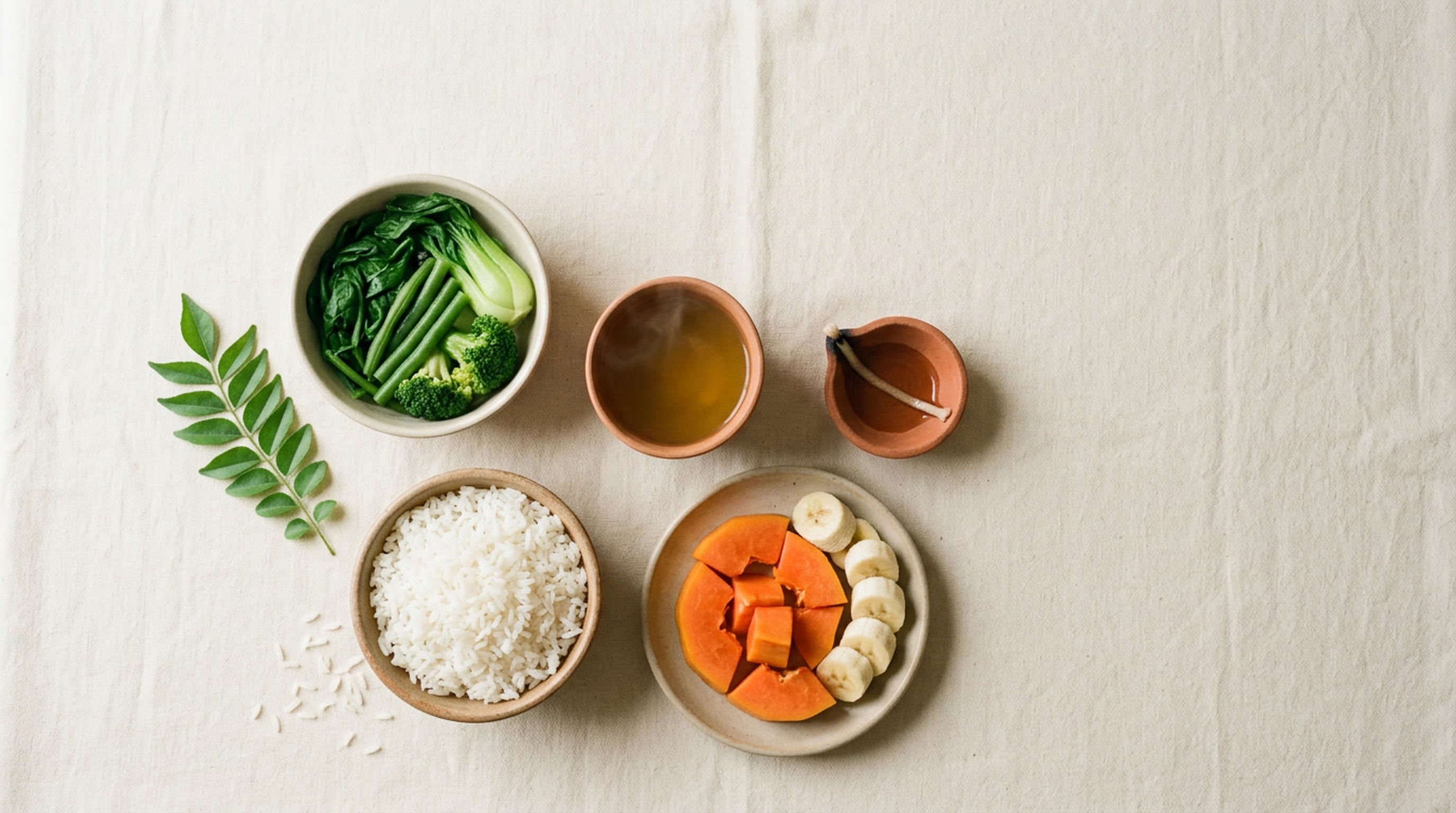 An overhead flat-lay of a simple light meal for a quiet Vesak eating reset: a bowl of rice, steamed greens, fresh papaya and banana, a cup of golden herbal tea, a small clay oil lamp, and a sprig of curry leaves on cream linen.