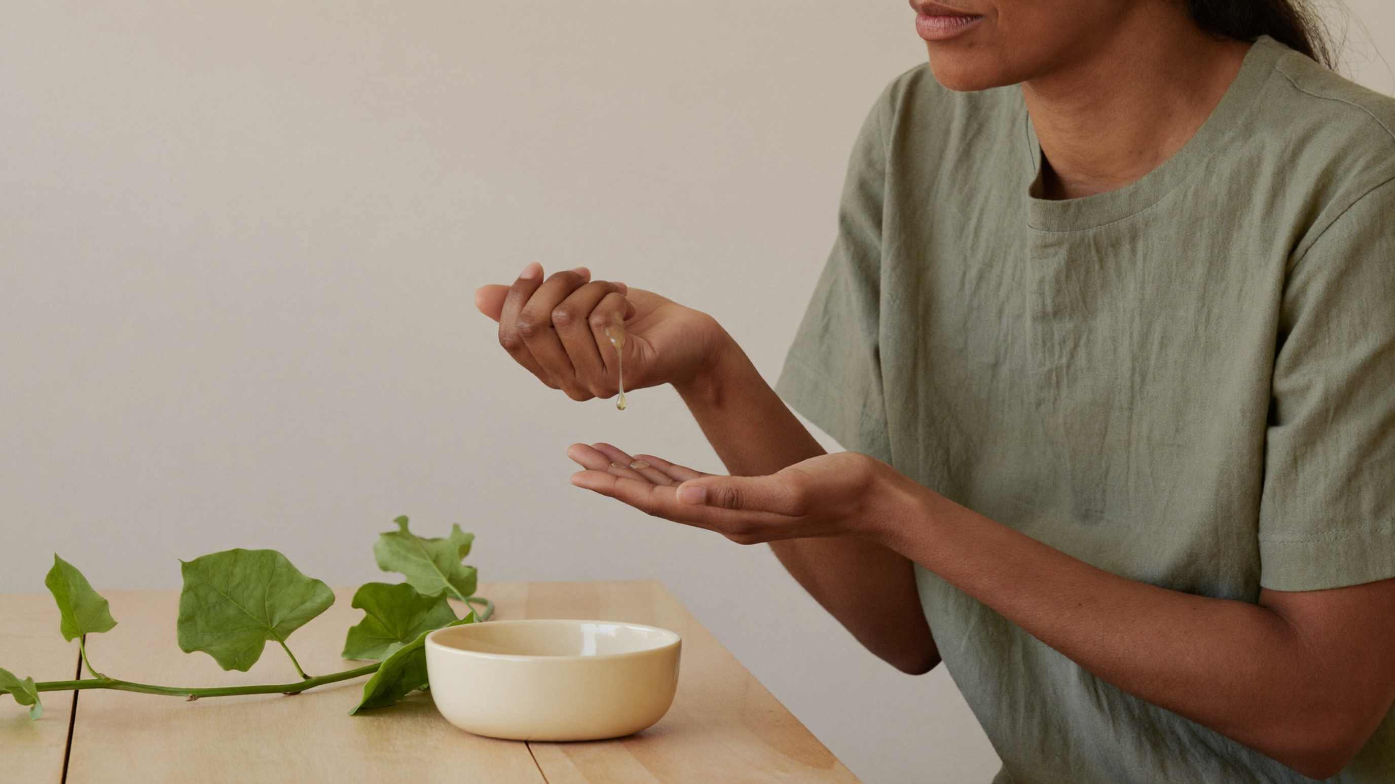 A South Asian woman pressing a drop of light facial oil into her palm at a sunlit wooden table, with a sprig of fresh green welpenela leaves and a plain cream ceramic bowl beside her.