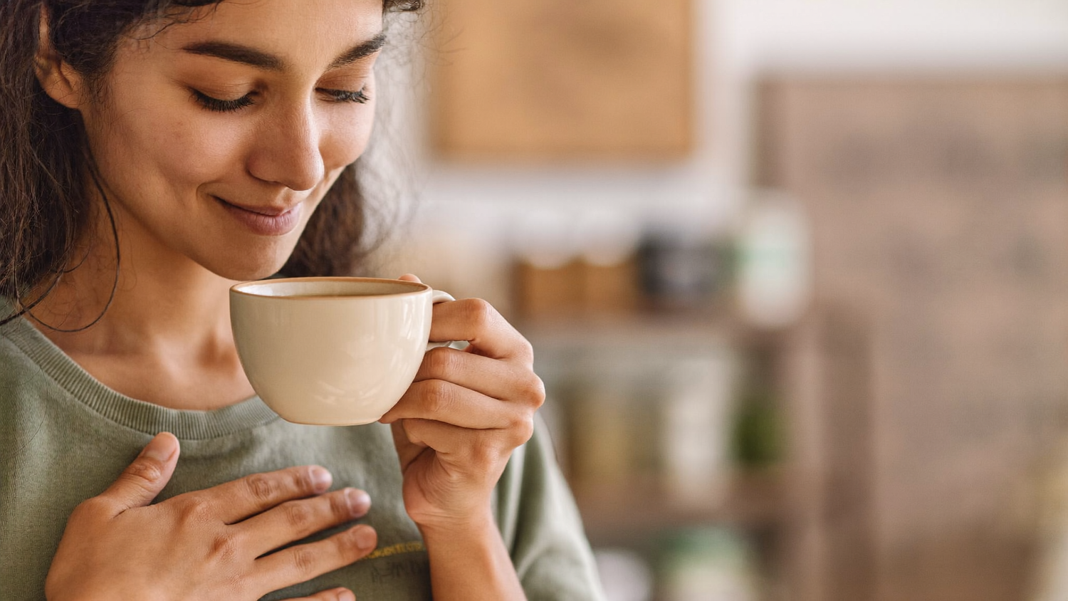 A South Asian woman in her early thirties holding a plain ceramic cup of warm herbal tea with both hands near her chest in soft morning light.