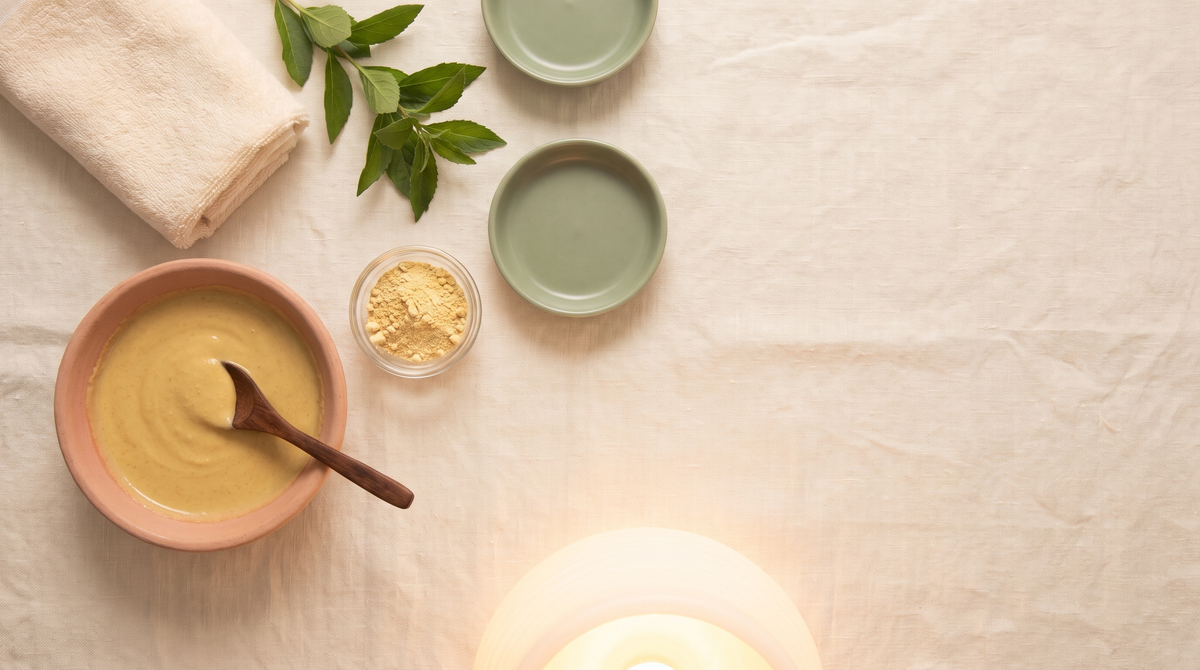 Flat-lay of an evening face-pack ritual on cream linen: a clay bowl of smooth golden herbal paste with a wooden spoon, a small glass dish of pale yellow root powder, a folded cotton towel, green leaves and a softly glowing lamp