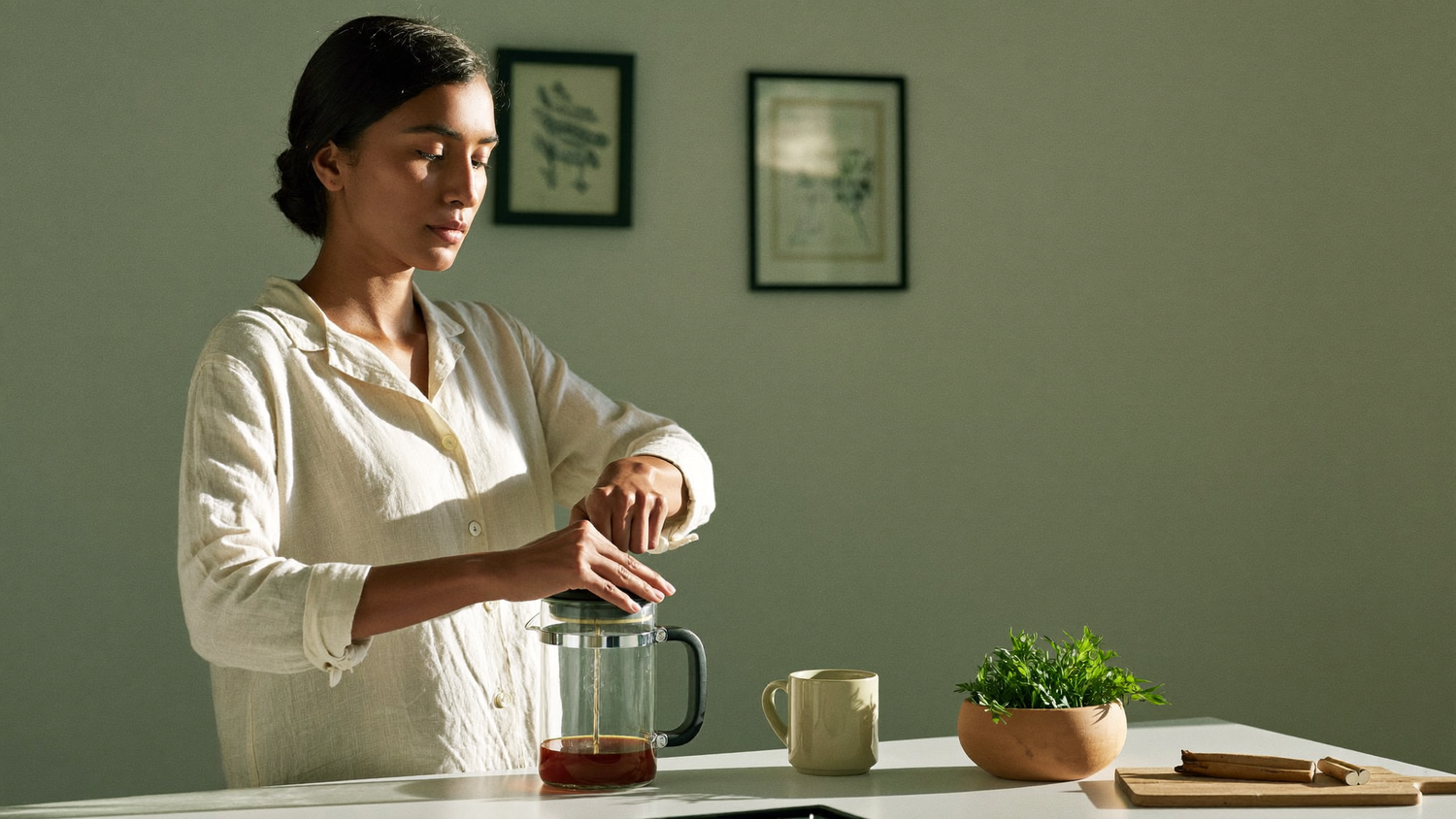A South Asian woman pressing fresh coffee in a glass French press at a sunlit kitchen counter on a calm weekend morning, with a bowl of green herb leaves and cinnamon quills nearby.