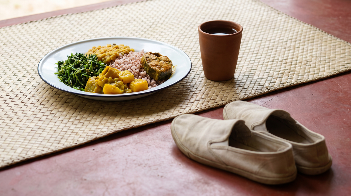 A Sri Lankan rice and curry plate with mallung, pumpkin curry, red rice, dhal and fish, beside a terracotta cup of tea and plain canvas shoes on a red veranda floor