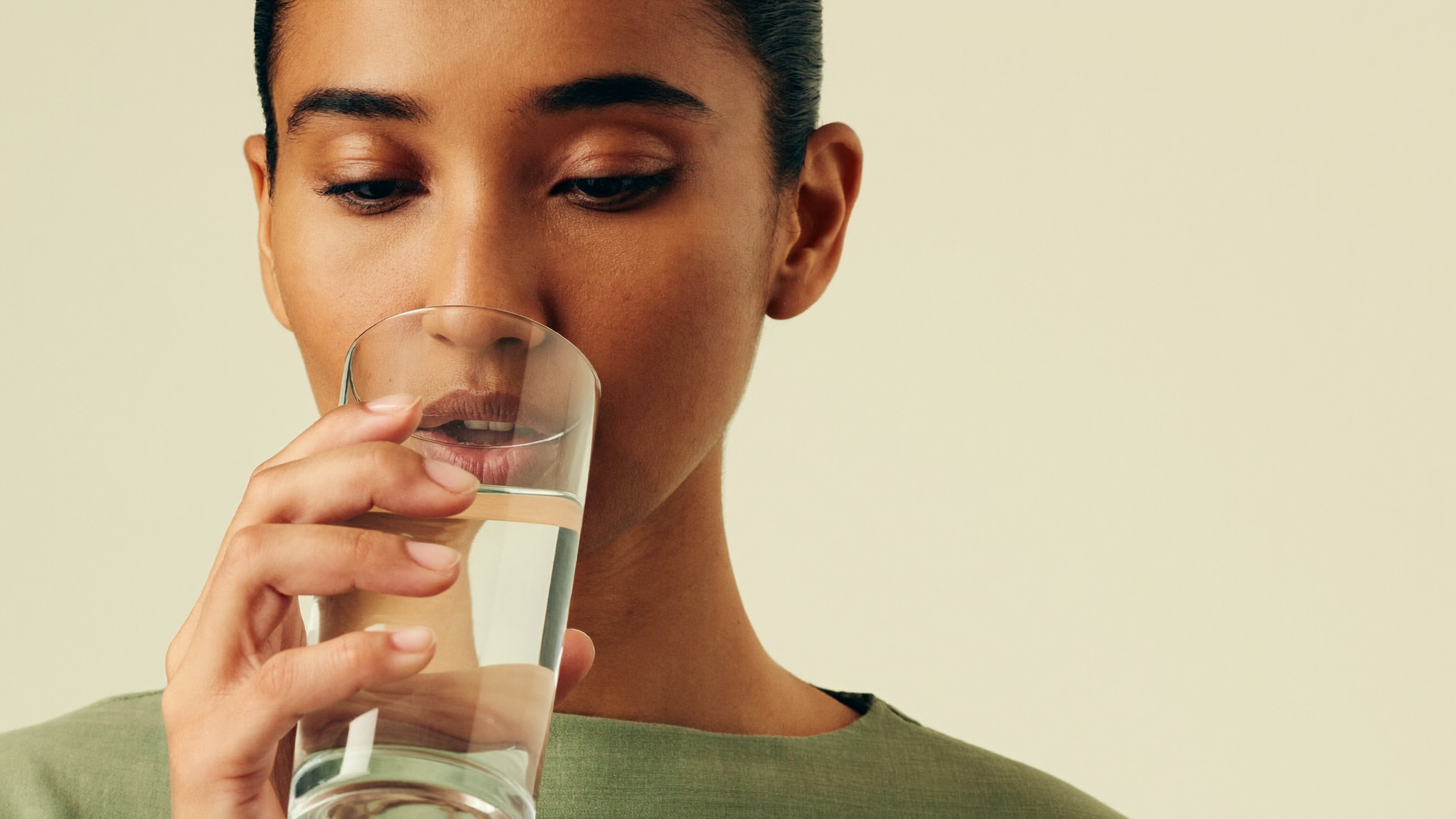 A South Asian woman drinking a tall clear glass of water against a plain cream background in soft natural light.